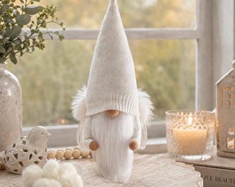 May include: A white gnome figurine with a long, pointed hat and a fluffy white beard. The gnome has a knitted hat and a wooden nose and hands. The figurine is displayed on a wooden surface, with a candle and decorative items in the background.