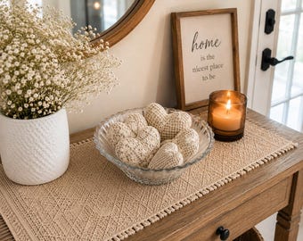 Puede incluir: Un cuenco de cristal transparente lleno de varios adornos en forma de corazón de ganchillo beige. Junto al cuenco hay un jarrón blanco texturizado con flores de paniculata, una vela encendida en un tarro de cristal marrón y un cartel de madera que dice Home is the nicest place to be.