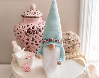 May include: A decorative gnome with a light blue knit hat and matching sleeves. The hat is adorned with a floral garland. The gnome has a long white beard, a tan nose, and small orange hands. It is a whimsical home decor item.