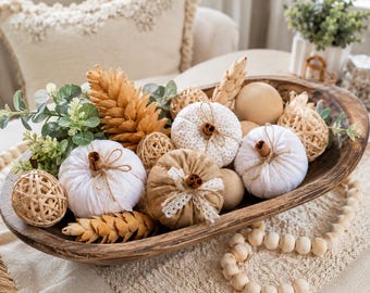 May include: A dark brown wooden dough bowl filled with fabric pumpkins in white and tan, woven rattan spheres, pinecones, and green foliage. The pumpkins feature cinnamon stick stems and lace accents, arranged alongside wooden beads on a textured fabric.