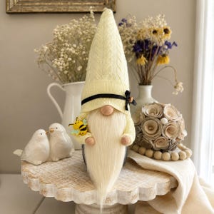 May include: A whimsical gnome figurine with a long, cream-colored beard and a yellow knit hat. The gnome is holding a small, yellow and black bee. A black ribbon with a bow accents the hat. Decorative birds and flowers are in the background.