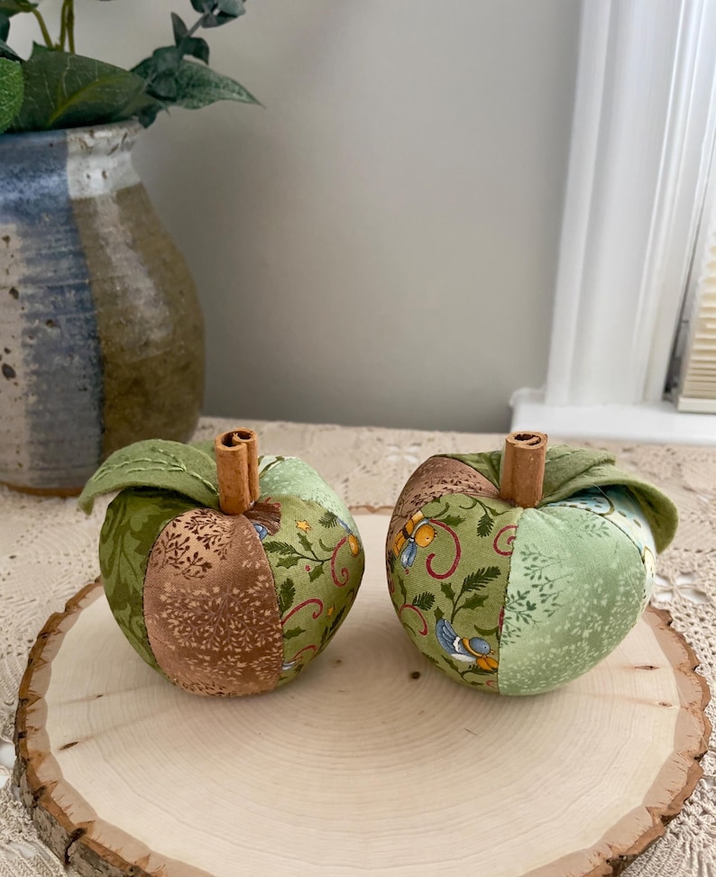 Puede incluir: Dos manzanas de tela hechas de segmentos de patchwork de tela de algod&oacute;n con patrones verdes y marrones. Cada manzana presenta una hoja de tela verde y un tallo de rama de canela que sobresale del centro superior de la fruta.