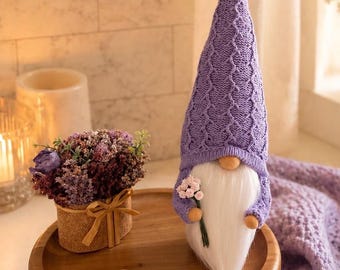 May include: A decorative gnome with a long white beard and a lavender knit hat and sweater. The gnome is holding a small bouquet of pink flowers. It is sitting on a wooden tray with a small potted plant and two wooden hearts.