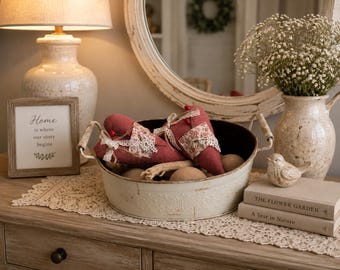 May include: A white metal oval basin with two handles contains two red fabric hearts with white lace and floral trim, alongside wooden spheres. The basin sits on a lace runner next to a framed sign reading Home is where our story begins.