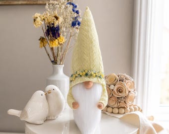 May include: A decorative gnome with a yellow knit hat and a long white beard. The hat is adorned with a floral band. Two white ceramic bird figurines and a vase of dried flowers are also present.