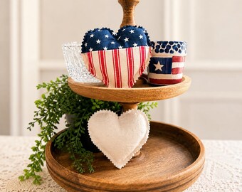 May include: A two-tiered wooden tray holds patriotic-themed decorations. The top tier displays a clear glass bowl, two heart-shaped pillows with red and white stripes and blue stars, and a small ceramic cup with a flag design.