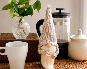 May include: A decorative gnome figurine featuring a tall, textured beige knit hat, a long white faux fur beard, and a small white mug held in its hands with the word COFFEE printed in black text on the front of the miniature mug.
