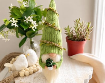 May include: A decorative gnome figurine with a green knit hat adorned with faux vines and leaves. The gnome has a long white beard and is holding a small potted plant. The figurine is displayed on a white, scalloped-edge stand.
