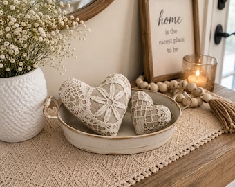 May include: Two beige fabric hearts decorated with white lace floral patterns rest inside a shallow, oval, white metal tray with side handles. The tray sits on a textured beige runner next to a white vase and a wooden sign reading Home is the nicest place to be.