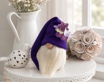 May include: A gnome figurine featuring a long, cream-colored faux fur beard, a round nose, and a tall, pointed purple fabric hat adorned with a cluster of small, light and dark purple hydrangea-like silk flowers attached to the side.