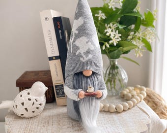 May include: A decorative gnome figurine with a gray knit hat and beard, holding a miniature tray with a cup of coffee. The gnome is standing on a white, distressed wooden surface, with books and a bird figurine in the background.