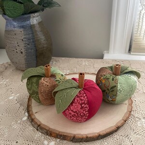Puede incluir: Tres manzanas de tela rellenas con dise&ntilde;os de patchwork en tonos verde, marr&oacute;n y rojo. Cada manzana tiene un tallo de rama de canela y hojas de tela verde con detalles bordados. Las manzanas est&aacute;n dispuestas sobre una rodaja de madera.
