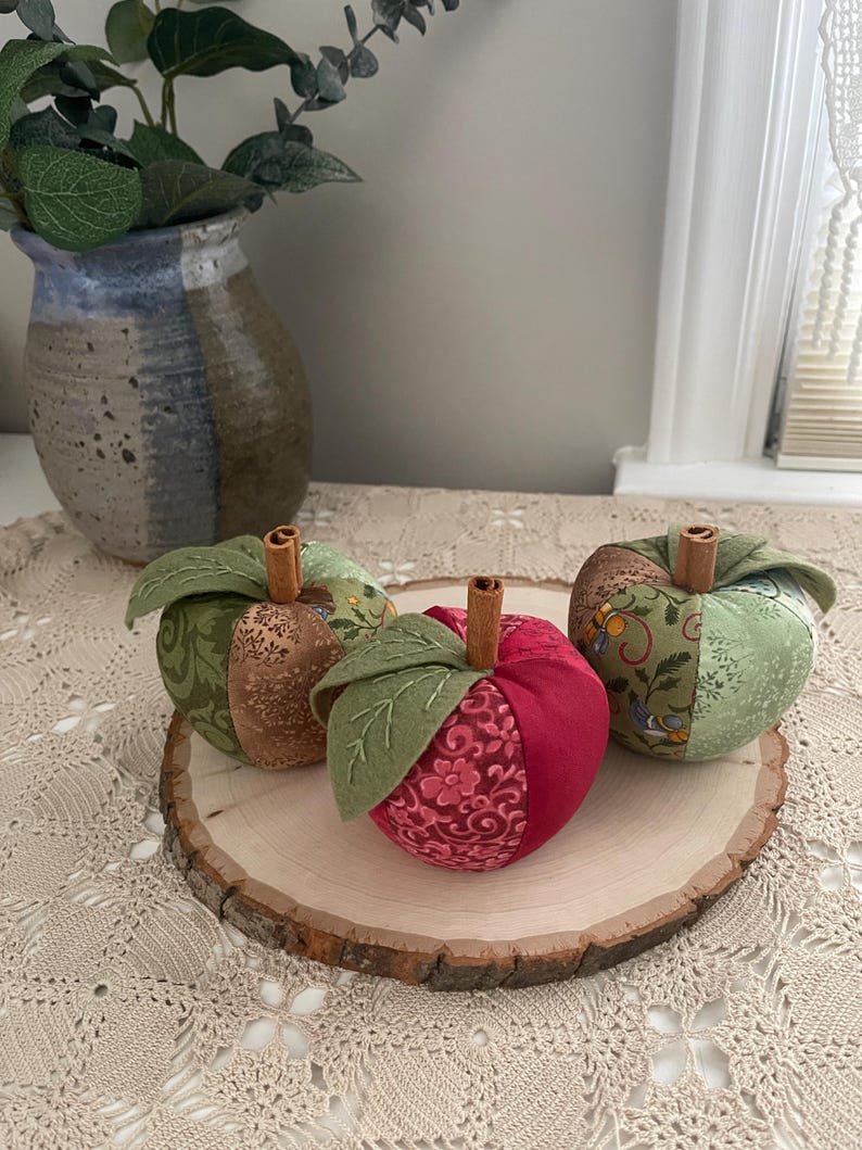 Puede incluir: Tres manzanas de tela rellenas con dise&ntilde;os de patchwork en tonos verde, marr&oacute;n y rojo. Cada manzana tiene un tallo de rama de canela y hojas de tela verde con detalles bordados. Las manzanas est&aacute;n dispuestas sobre una rodaja de madera.