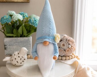 May include: A light blue gnome figurine with a long white beard, wearing a matching knit hat and sweater. The gnome has a small yellow flower on its hat and a round orange nose. The figurine is on a white cake stand.
