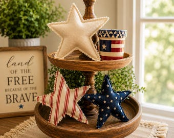 May include: A wooden tiered tray displays patriotic decor. It features a white star, a red and white striped star, and a blue star with white stars. A small ceramic cup with an American flag design is also present.