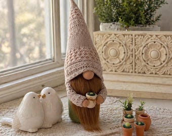 Puede incluir: Una figura decorativa de gnomo con una larga barba marrón y un gorro de punto. Dos figuras de pájaros de cerámica blanca están junto al gnomo. También se ven pequeñas plantas en macetas y recortes de madera en forma de corazón.