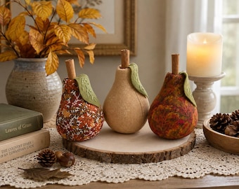 May include: Three pear-shaped ornaments made of fabric, each topped with a cinnamon stick stem and a green felt leaf. Two pears feature a multi-colored autumn leaf print in shades of orange, red, and brown, while the center pear is solid tan.