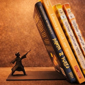 Könnte beinhalten: Ein dekoratives Buchstützen-Set mit einer kleinen Figur eines Zauberers, der einen Zauberstab hält und einen Stapel farbiger polnischer Bücher stützt. Die Buchstütze steht auf einer braunen Oberfläche.