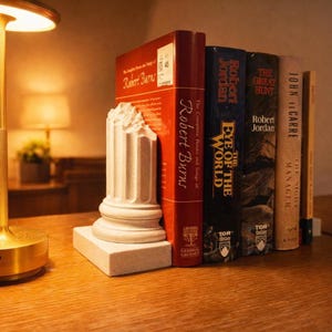 Puede incluir: Un sujetalibros de columna clásica blanca sostiene una fila de libros sobre una superficie de madera. Los libros tienen títulos como "Robert Burns" y "The Eye of the World". Una lámpara dorada es visible en el fondo.