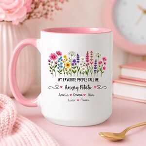May include: White ceramic mug with a pink handle and interior. The mug features a floral design with the text "MY FAVORITE PEOPLE CALL ME Angry Potato" and names below. A gold spoon rests on a pink surface.