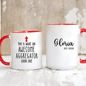 May include: Two white ceramic mugs with red handles and rims. One mug reads "THIS IS WHAT AN AWESOME AGGREGATOR LOOKS LIKE" with an arrow. The other mug says "Gloria EST 2026" in black script.