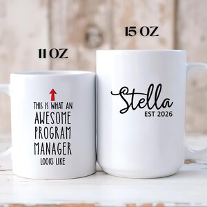 Puede incluir: Dos tazas de cer&aacute;mica blancas. La taza m&aacute;s peque&ntilde;a, 312 ml, tiene el texto "THIS IS WHAT AN AWESOME PROGRAM MANAGER LOOKS LIKE" con una flecha roja. La taza m&aacute;s grande, 425 ml, tiene el nombre "Stella" y "EST 2026" en escritura negra.