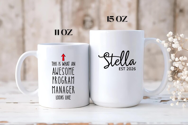 Puede incluir: Dos tazas de cer&aacute;mica blancas. La taza m&aacute;s peque&ntilde;a, 312 ml, tiene el texto "THIS IS WHAT AN AWESOME PROGRAM MANAGER LOOKS LIKE" con una flecha roja. La taza m&aacute;s grande, 425 ml, tiene el nombre "Stella" y "EST 2026" en escritura negra.