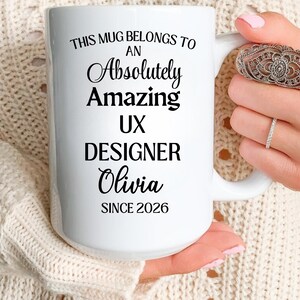 Può includere: Tazza in ceramica bianca con testo nero: "THIS MUG BELONGS TO AN Absolutely Amazing UX DESIGNER Olivia SINCE 2026". La tazza ha un manico grande. La tazza è tenuta da una persona.