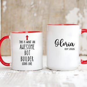Op de afbeelding: Twee witte keramische mokken met rode handvatten en randen. Op één mok staat "THIS IS WHAT AN AWESOME BOT BUILDER LOOKS LIKE" met een pijl. De andere mok zegt "Gloria EST 2026".