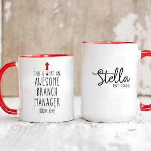 May include: Two white ceramic mugs with red interiors and handles. One mug reads "THIS IS WHAT AN AWESOME BRANCH MANAGER LOOKS LIKE" with an arrow. The other mug has the name "Stella" and "EST 2026".
