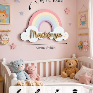 May include: A wall-mounted rainbow decoration featuring a white cloud base with the custom name Mackenzie in gold script lettering. The rainbow has pastel stripes of pink, yellow, blue, and purple. The piece measures 50cm in width.