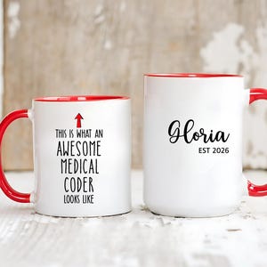 Peut inclure: Deux mugs en céramique blanche avec des poignées et des bords rouges. L'un des mugs indique "This is what an AWESOME MEDICAL CODER looks like" avec une flèche. L'autre mug dit "Gloria EST 2026".