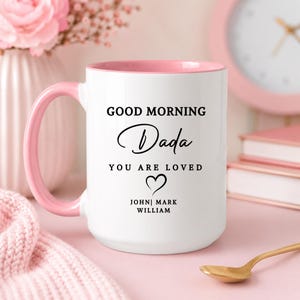 May include: White ceramic mug with a pink interior and handle. The mug features the text "GOOD MORNING Dada YOU ARE LOVED" with a heart symbol and names below. A gold spoon rests on the table.