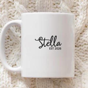 Puede incluir: Taza de cer&aacute;mica blanca con asa curva. La taza presenta el nombre "Stella" en fuente cursiva, con "EST 2026" impreso debajo en una fuente m&aacute;s peque&ntilde;a, sin serifa. La taza est&aacute; sobre un fondo texturizado blanquecino.