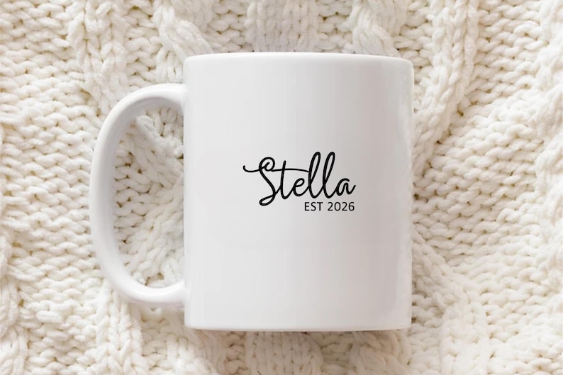 Puede incluir: Taza de cer&aacute;mica blanca con asa curva. La taza presenta el nombre "Stella" en fuente cursiva, con "EST 2026" impreso debajo en una fuente m&aacute;s peque&ntilde;a, sin serifa. La taza est&aacute; sobre un fondo texturizado blanquecino.