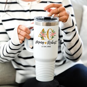 May include: White travel mug with a handle, featuring the text "40" in gold with floral accents, the names "Mary & Robert", and the date "17.04.1984". The mug has a silver lid and a clear straw.