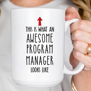 Puede incluir: Taza de cer&aacute;mica blanca con el texto "THIS IS WHAT AN AWESOME PROGRAM MANAGER LOOKS LIKE" en fuente negra. Una flecha roja apunta hacia arriba. La taza es sostenida por una persona.