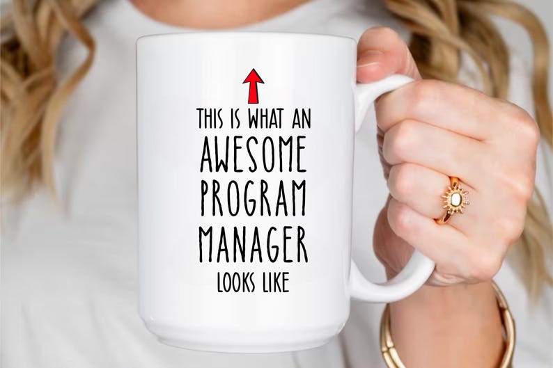 Puede incluir: Taza de cer&aacute;mica blanca con el texto "THIS IS WHAT AN AWESOME PROGRAM MANAGER LOOKS LIKE" en fuente negra. Una flecha roja apunta hacia arriba. La taza es sostenida por una persona.