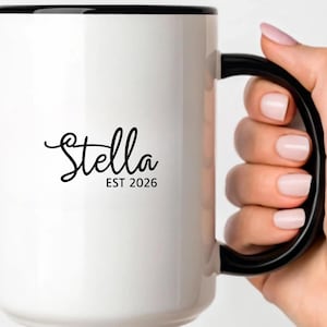 Puede incluir: Taza de cer&aacute;mica blanca con borde y asa negros. La taza presenta el nombre "Stella" en cursiva, con "EST 2026" debajo. La taza est&aacute; sostenida por una persona.