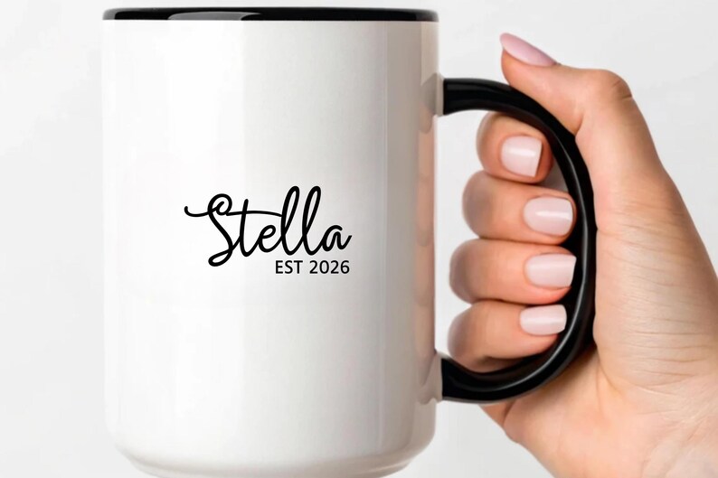 Puede incluir: Taza de cer&aacute;mica blanca con borde y asa negros. La taza presenta el nombre "Stella" en cursiva, con "EST 2026" debajo. La taza est&aacute; sostenida por una persona.