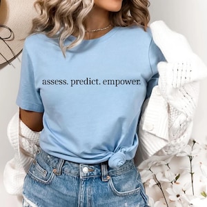 May include: A pale blue t-shirt with the text "assess. predict. empower." in black. The shirt is knotted at the waist and worn with denim shorts. A white cardigan is draped over the shoulders. A straw hat is visible in the background.