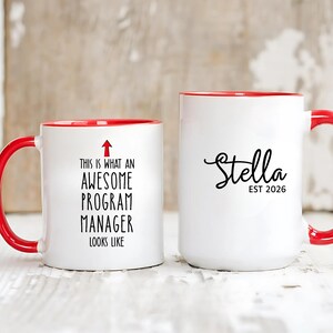 May include: Two white ceramic mugs with red handles and rims. One mug has the text "THIS IS WHAT AN AWESOME PROGRAM MANAGER LOOKS LIKE" with an arrow. The other mug says "Stella EST 2026".