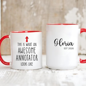 Könnte beinhalten: Zwei weiße Keramikbecher mit roten Griffen und Rändern. Auf einem Becher steht "THIS IS WHAT AN AWESOME ANNOTATOR LOOKS LIKE" mit einem Pfeil. Der andere Becher hat den Namen "Gloria" und "EST 2026" in schwarzer Schrift.