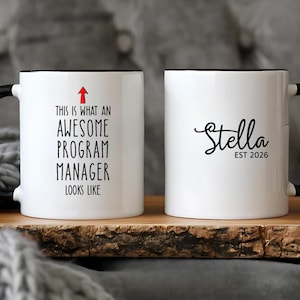 Puede incluir: Taza de cer&aacute;mica blanca con asa y borde negros. Un lado dice "THIS IS WHAT AN AWESOME PROGRAM MANAGER LOOKS LIKE" con una flecha roja. El otro lado dice "Stella EST 2026" en escritura negra.