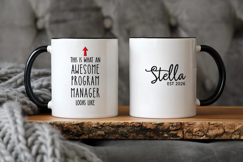 Puede incluir: Taza de cer&aacute;mica blanca con asa y borde negros. Un lado dice "THIS IS WHAT AN AWESOME PROGRAM MANAGER LOOKS LIKE" con una flecha roja. El otro lado dice "Stella EST 2026" en escritura negra.