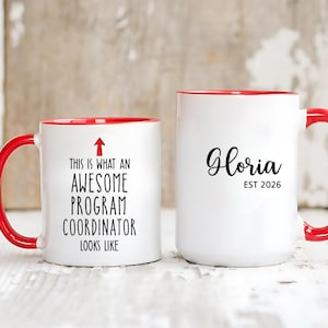 May include: Two white ceramic mugs with red handles and rims. One mug reads "THIS IS WHAT AN AWESOME PROGRAM COORDINATOR LOOKS LIKE" with an arrow. The other mug says "Gloria EST 2026".