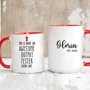 May include: Two white ceramic mugs with red handles and rims. One mug reads "This is what an AWESOME OUTPUT TESTER looks like" with an arrow. The other mug says "Gloria EST 2026" in black script.