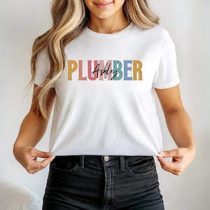 Puede incluir: Camiseta blanca con la palabra "PLUMBER" en letras de bloque coloridas en el pecho. El nombre "Ashley" está escrito en cursiva entre el texto "PLUMBER". La camiseta está hecha de un material suave.