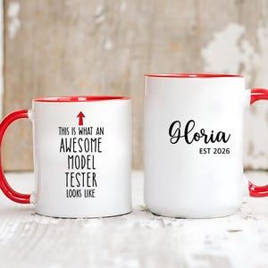 Può includere: Due tazze in ceramica bianca con manici e bordi rossi. Una tazza recita "THIS IS WHAT AN AWESOME MODEL TESTER LOOKS LIKE" con una freccia. L'altra tazza dice "Gloria EST 2026" in corsivo nero.