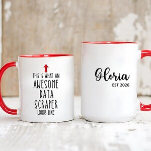 Puede incluir: Dos tazas de cerámica blanca con asas y bordes rojos. Una taza dice "This is what an AWESOME DATA SCRAPER looks like" con una flecha. La otra taza dice "Gloria EST 2026" en escritura negra.